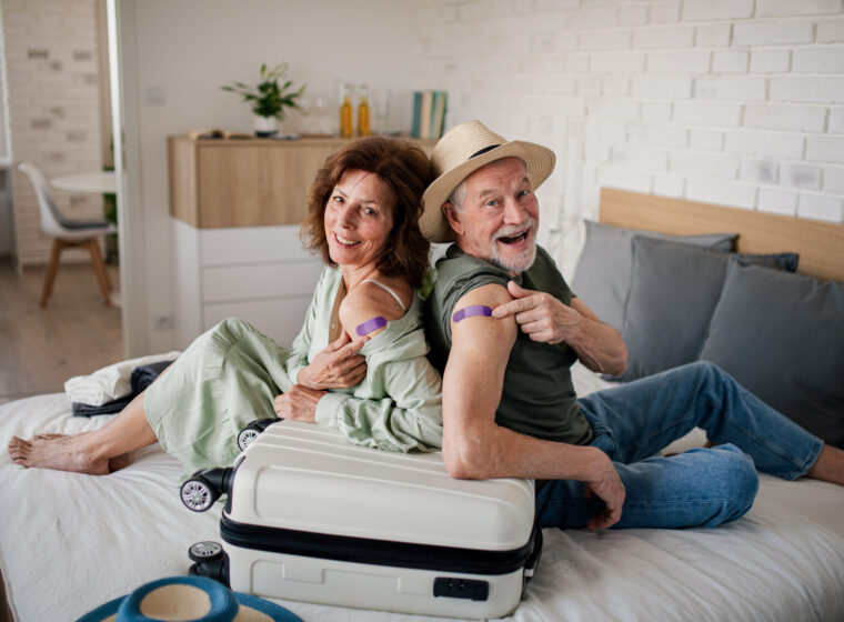 Senior couple sitting on bed indoors in hotel room, coronavirus vaccination, holiday and new normal concept.